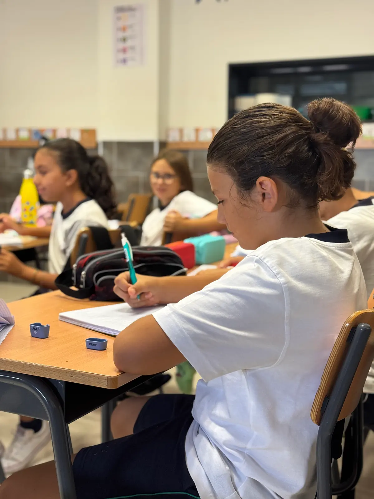 Alumna escribiendo en su cuaderno con sus compañeras de clase al fondo