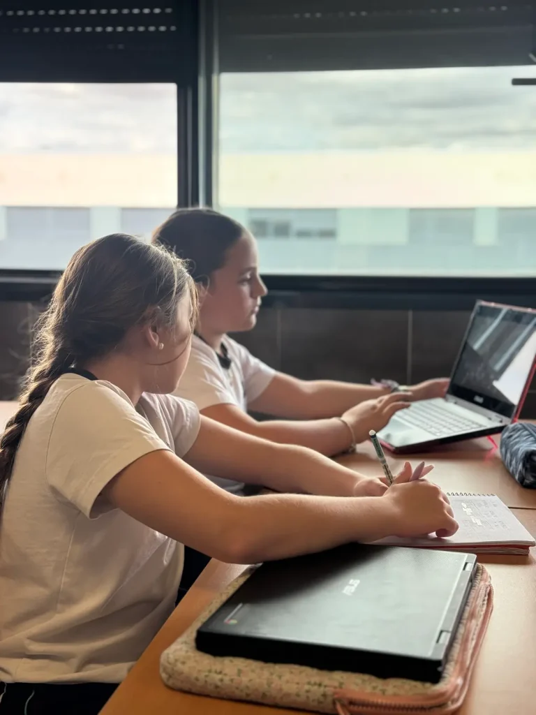Dos alumnas trabajando con ordenadores portátiles y cuaderno junto a la ventana del aula