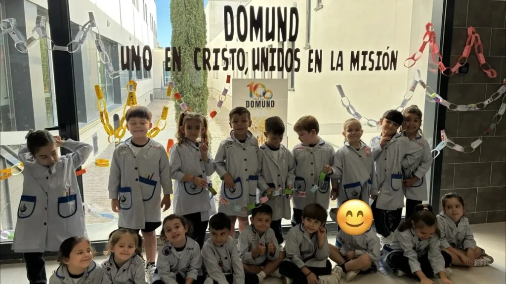 Grupo de alumnos con babi ante la cristalera del colegio decorada con cadenas de papel y el lema «DOMUND: Uno en Cristo, unidos en la misión»