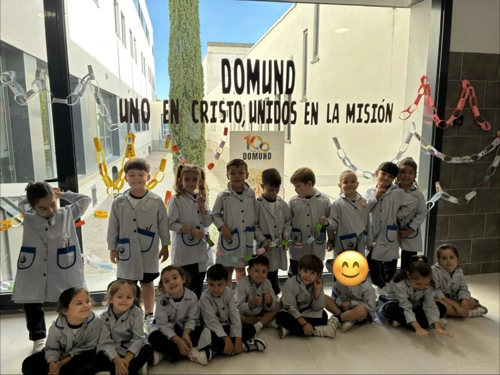 Grupo de alumnos sosteniendo eslabones de papel ante la cristalera decorada con el lema «Uno en Cristo, unidos en la misión» y el cartel de los 100 años del DOMUND
